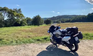 Biały motocykl turystyczny zaparkowany na drodze gruntowej, w tle pole i zalesione wzgórza.