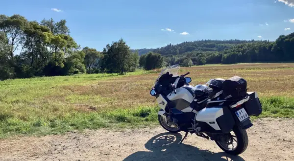 Biały motocykl turystyczny zaparkowany na drodze gruntowej, w tle pole i zalesione wzgórza.